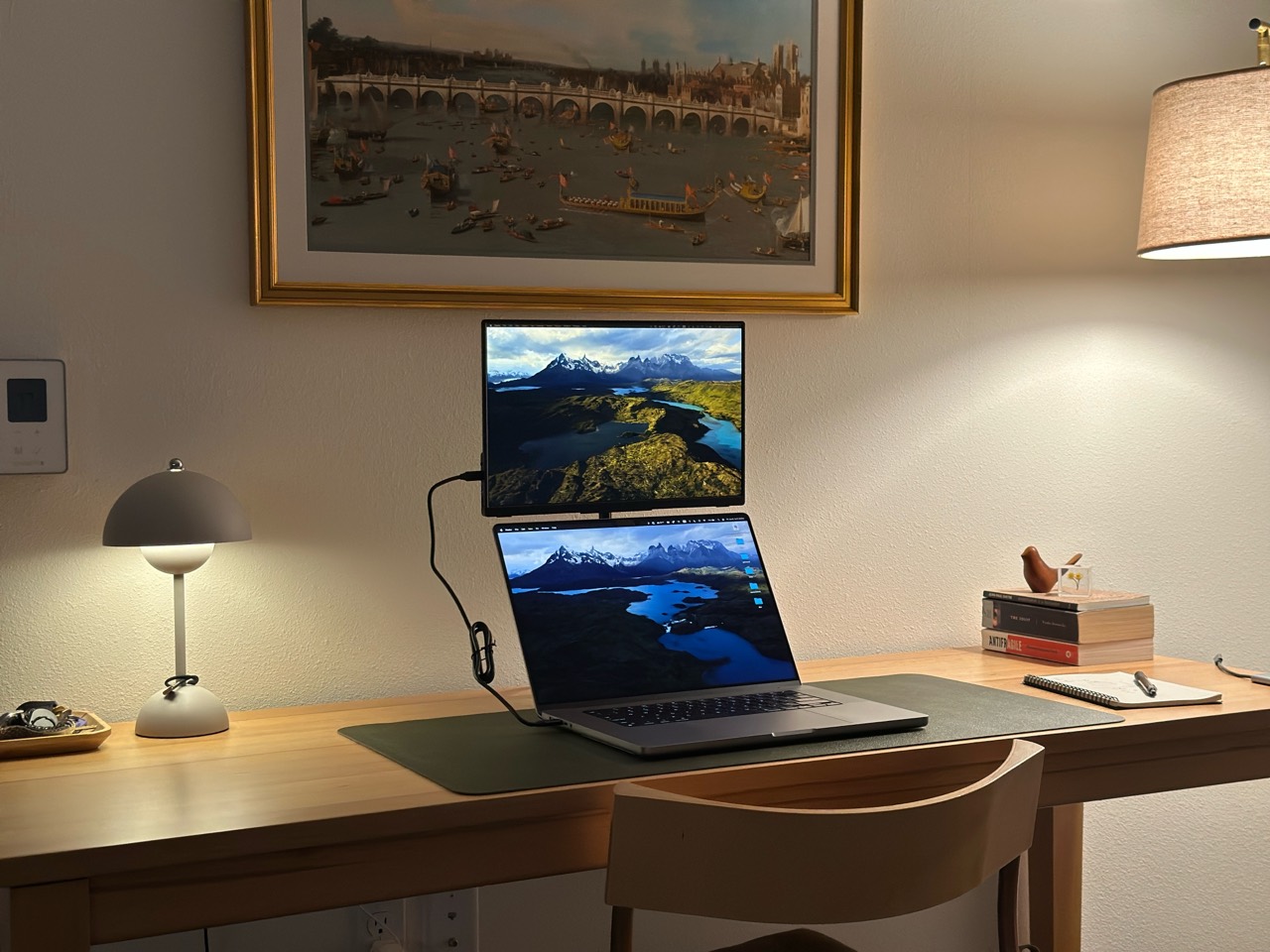 A warmly lit desk with a laptop, monitor, books, and lamps.